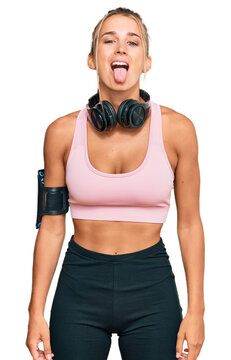 Young blonde woman wearing gym clothes and using headphones sticking tongue out happy with funny expression. emotion concept.