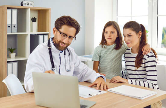 Friendly family doctor talking to mother and child. Man pediatrician in white medical coat sitting at desk together with mum and daughter and showing diagnostic test results on modern laptop computer