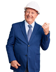 Senior handsome grey-haired man wearing suit and architect hardhat smiling with happy face looking and pointing to the side with thumb up.