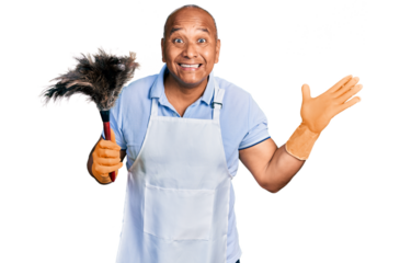 Hispanic middle age man wearing apron holding cleaning duster celebrating victory with happy smile and winner expression with raised hands