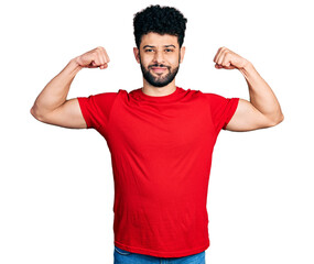 Young arab man with beard wearing casual red t shirt showing arms muscles smiling proud. fitness concept.
