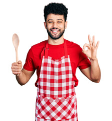 Young arab man with beard wearing baker uniform holding wooden spoon smiling and laughing hard out...
