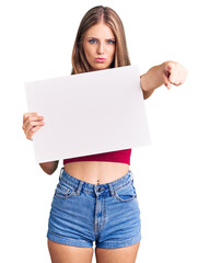 Fototapeta premium Young beautiful blonde woman holding blank empty banner pointing with finger to the camera and to you, confident gesture looking serious