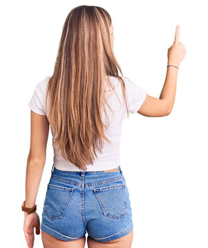Young beautiful blonde woman wearing casual white tshirt posing backwards pointing ahead with finger hand