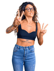 Young hispanic woman with tattoo wearing casual clothes and glasses smiling looking to the camera...