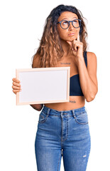 Young hispanic woman with tattoo holding empty white chalkboard serious face thinking about question with hand on chin, thoughtful about confusing idea