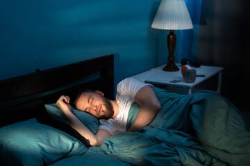 Handsome caucasian man sleeping cozily on a bed in his bedroom at night with blue nightly colors in...