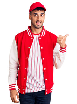 Young hispanic man wearing baseball uniform smiling with happy face looking and pointing to the side with thumb up.