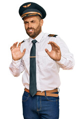 Handsome man with beard wearing airplane pilot uniform disgusted expression, displeased and fearful doing disgust face because aversion reaction. with hands raised