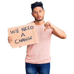 Handsome latin american young man holding we need a change banner with angry face, negative sign...