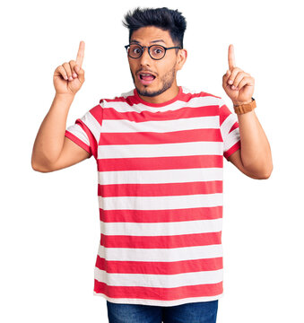 Handsome latin american young man wearing casual clothes and glasses smiling amazed and surprised and pointing up with fingers and raised arms.