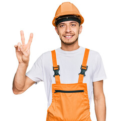 Hispanic young man wearing handyman uniform and safety hardhat smiling with happy face winking at the camera doing victory sign. number two.