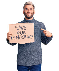 Young blond man holding save our democracy cardboard banner pointing finger to one self smiling happy and proud