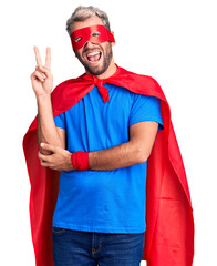 Young blond man wearing super hero custome smiling with happy face winking at the camera doing victory sign. number two.