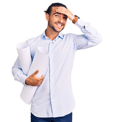Young handsome man holding paper blueprints stressed and frustrated with hand on head, surprised...