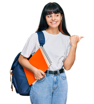 Young hispanic girl wearing student backpack and holding book pointing thumb up to the side smiling happy with open mouth