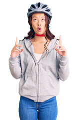 Young beautiful chinese girl wearing bike helmet amazed and surprised looking up and pointing with fingers and raised arms.