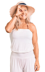 Middle age fit blonde woman wearing summer hat doing ok gesture with hand smiling, eye looking through fingers with happy face.