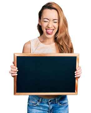 Young caucasian woman holding blackboard sticking tongue out happy with funny expression.