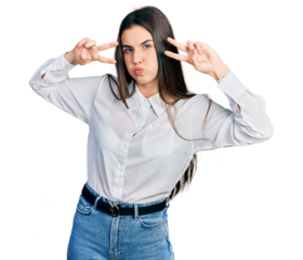 Young brunette teenager doing peace gesture close to eyes puffing cheeks with funny face. mouth inflated with air, catching air.