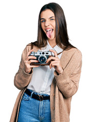 Young brunette teenager holding vintage camera sticking tongue out happy with funny expression.