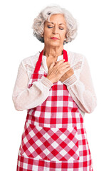 Senior grey-haired woman wearing apron smiling with hands on chest with closed eyes and grateful gesture on face. health concept.