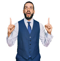 Young man with beard wearing business vest amazed and surprised looking up and pointing with fingers and raised arms.