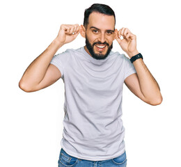 Young man with beard wearing casual white t shirt smiling pulling ears with fingers, funny gesture....