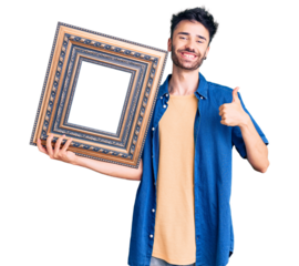 Young hispanic man holding empty frame smiling happy and positive, thumb up doing excellent and approval sign