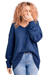 Young blonde woman with curly hair wearing casual winter sweater smelling something stinky and...