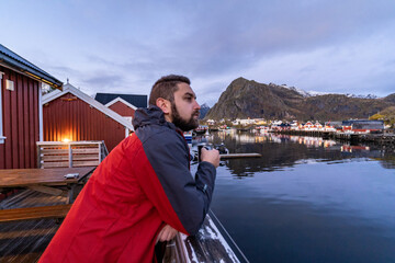 young man with beard drinking from the mug in the bay of Sørvågen typical norwegian village...