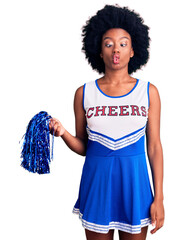 Young african american woman wearing cheerleader uniform holding pompom making fish face with lips,...