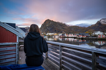 young woman drinking coffee in the morning and taking a look on the bay in Sørvågen fisherman village with red houses on the coast, lofoten islands, norway