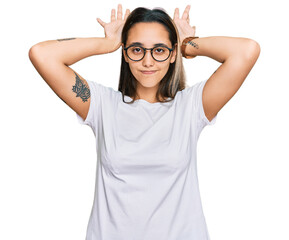 Young hispanic woman wearing casual white t shirt doing bunny ears gesture with hands palms looking...