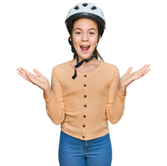 Beautiful brunette little girl wearing bike helmet celebrating victory with happy smile and winner expression with raised hands