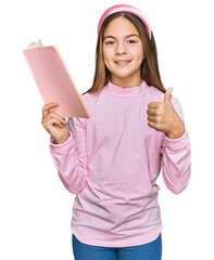 Beautiful brunette little girl reading a book smiling happy and positive, thumb up doing excellent and approval sign
