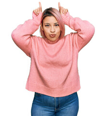 Hispanic woman with pink hair wearing casual winter sweater doing funny gesture with finger over head as bull horns