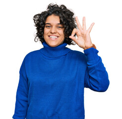 Young hispanic woman with curly hair wearing turtleneck sweater smiling positive doing ok sign with hand and fingers. successful expression.