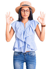Young beautiful latin girl wearing summer hat and glasses relax and smiling with eyes closed doing meditation gesture with fingers. yoga concept.
