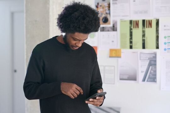 Focused black and afro man sending a text message in the office. Moodboard of varied business projects in the background