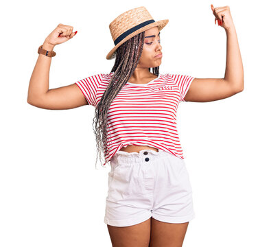 Young african american woman with braids wearing summer hat showing arms muscles smiling proud. fitness concept.