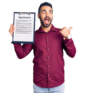 Young hispanic man holding clipboard with agreement document pointing thumb up to the side smiling happy with open mouth
