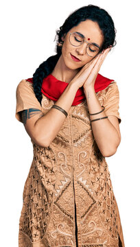 Young indian woman wearing traditional dress and glasses sleeping tired dreaming and posing with hands together while smiling with closed eyes.