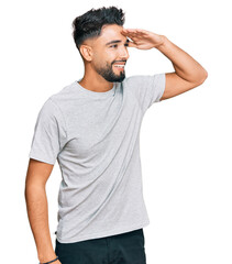 Young man with beard wearing casual grey tshirt very happy and smiling looking far away with hand over head. searching concept.