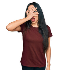 Young hispanic girl wearing casual t shirt peeking in shock covering face and eyes with hand, looking through fingers with embarrassed expression.