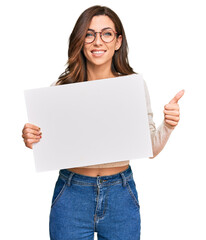 Young brunette woman holding blank empty banner smiling happy and positive, thumb up doing excellent and approval sign