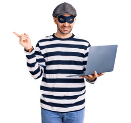 Young handsome man wearing burglar mask using laptop smiling happy pointing with hand and finger to...