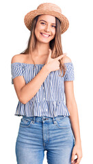 Young beautiful girl wearing hat and t shirt cheerful with a smile of face pointing with hand and finger up to the side with happy and natural expression on face