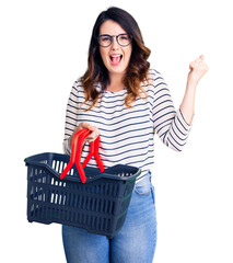 Beautiful young brunette woman holding supermarket shopping basket screaming proud, celebrating victory and success very excited with raised arms