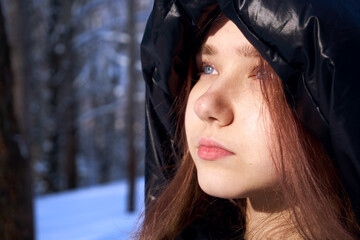 A teenage girl with blue, azure eyes looks to the side of her head with a hood draped over her head. Winter background